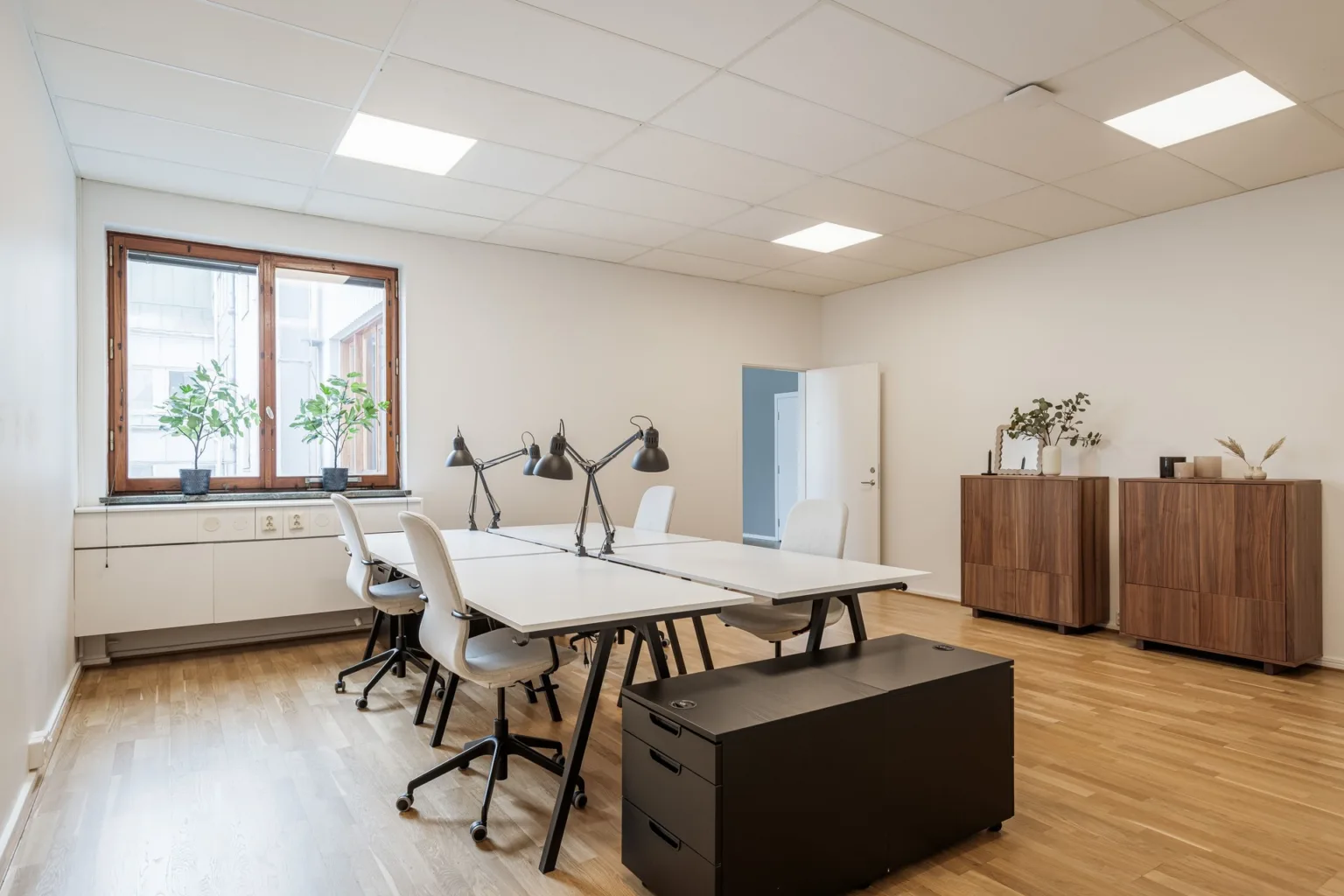 Ett kontorsutrymme med två stora skrivbord, stolar, skrivbordslampor och ett träskåp, belyst av naturligt ljus från ett fönster.
