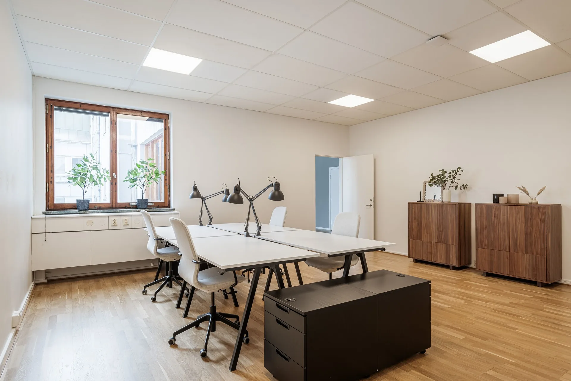Ett kontorsutrymme med två stora skrivbord, stolar, skrivbordslampor och ett träskåp, belyst av naturligt ljus från ett fönster.