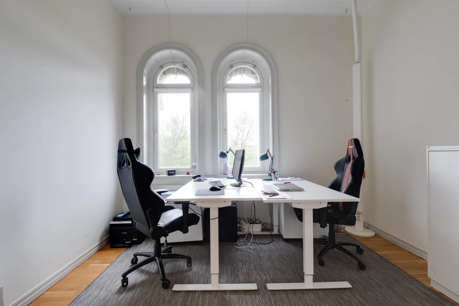 Ett arbetsrum med två skrivbord, svarta kontorsstolar och tre valvfönster.