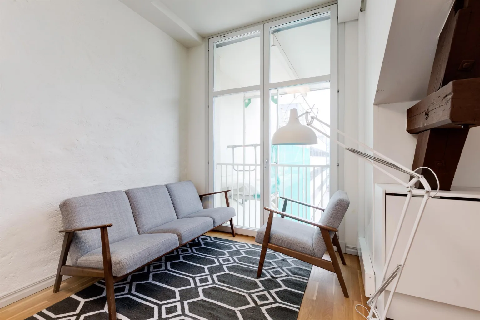Ett ljust rum med grå soffa och fåtöljer, geometrisk matta och stora fönster med balkongutsikt.