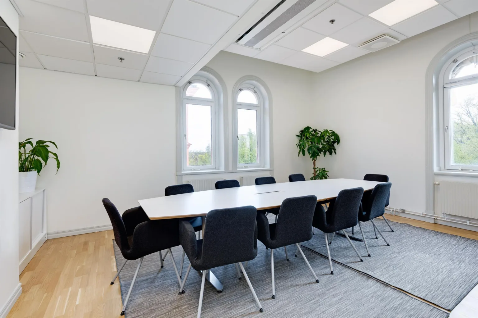 Ett konferensrum med ett stort vitt bord, svarta stolar, två stora fönster och en grön växt i hörnet.