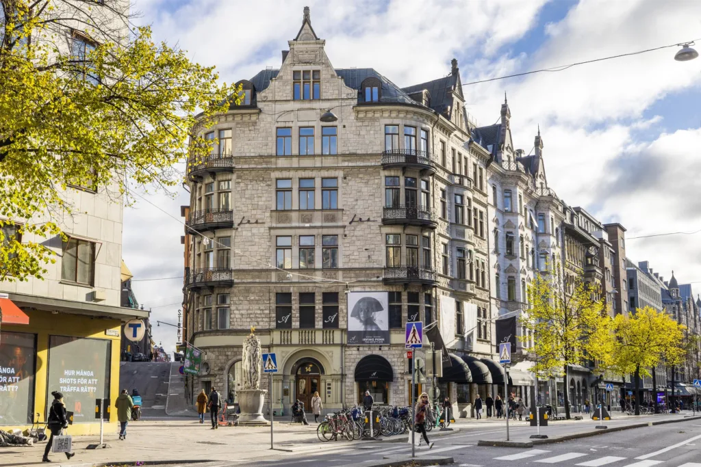 En stor stenbyggnad med flera våningar och utsmyckade detaljer, omgiven av träd och människor.