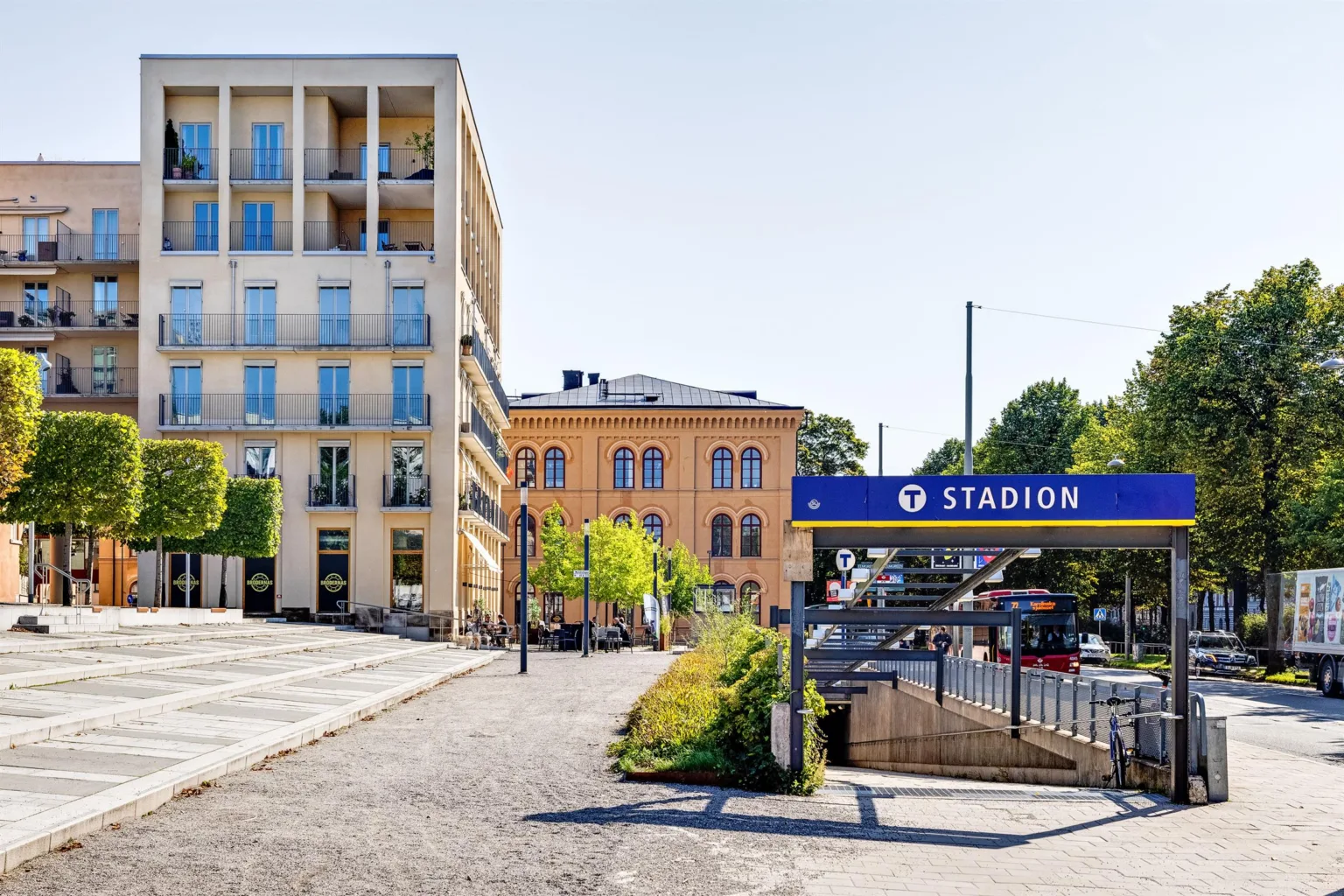 Gatumiljö med en skylt som visar "Stadion" vid tunnelbanestation, en modern byggnad och en gammal ljusgul byggnad i bakgrunden.