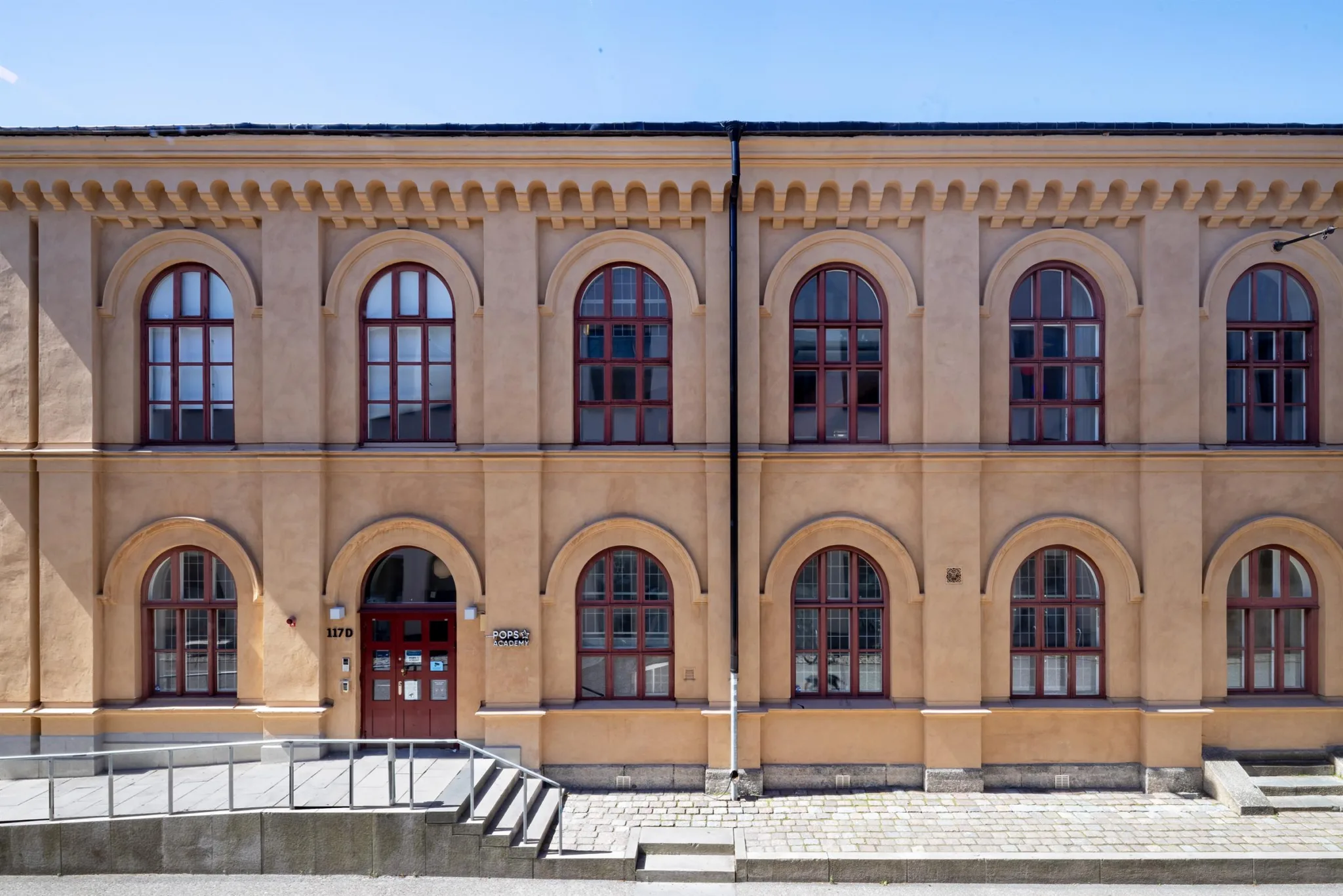 Närbild av en ljusgul byggnad med välvda fönster och röda dörrar, omgiven av stenläggning och en låg trappa.