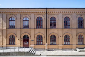 Närbild av en ljusgul byggnad med välvda fönster och röda dörrar, omgiven av stenläggning och en låg trappa.