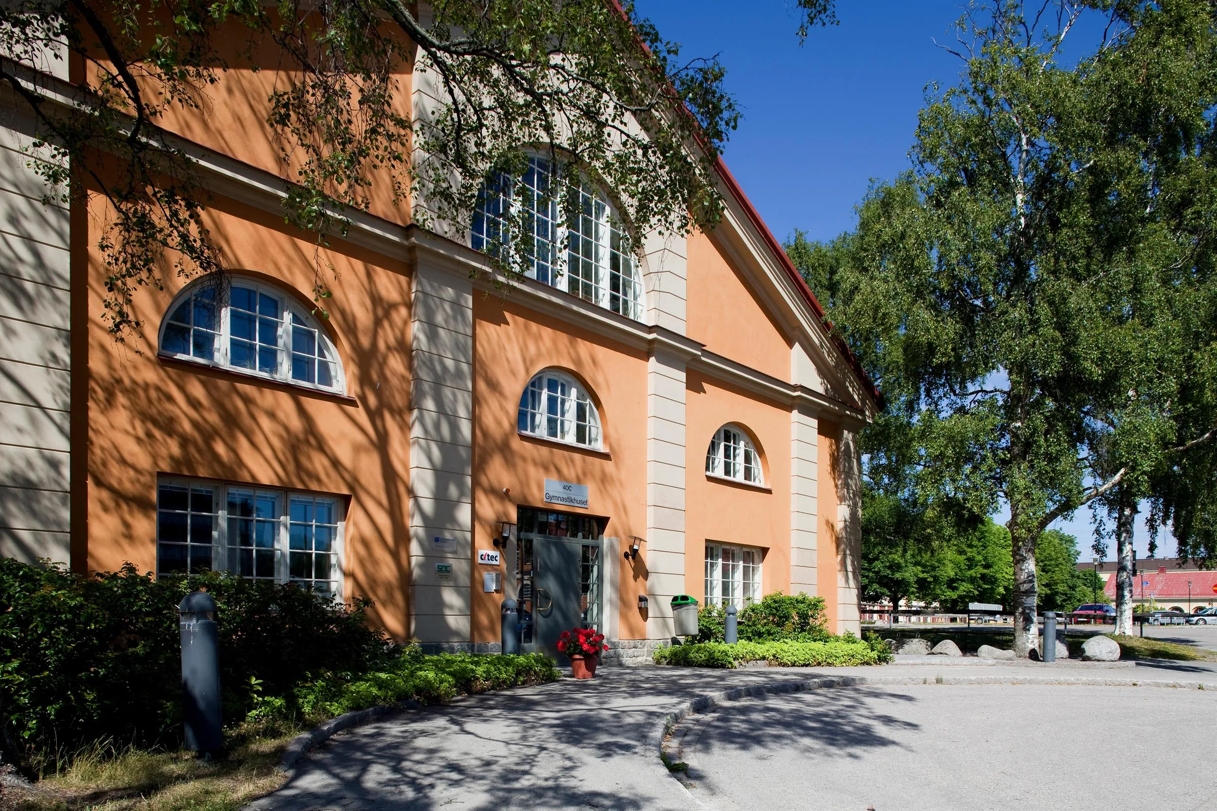 En orange byggnad med välvda fönster och detaljer i sten, omgiven av träd och en gångväg.