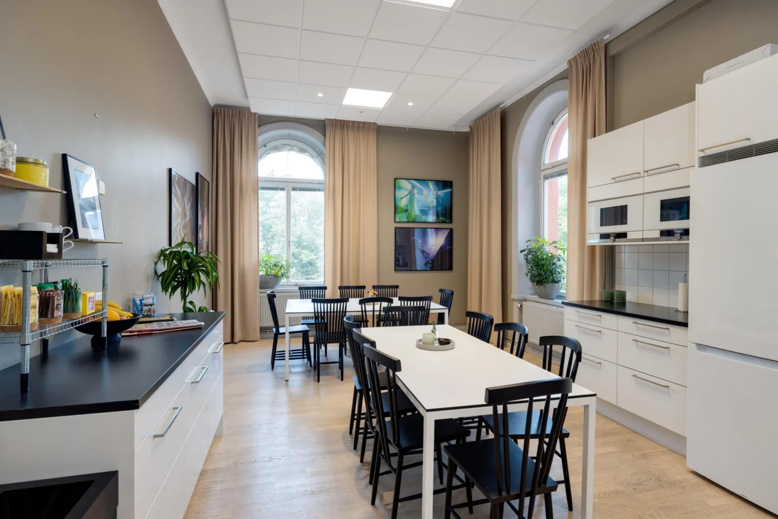Ett kök med ett långt bord omgivet av svarta stolar, vita skåp, växter och stora fönster med beige gardiner.