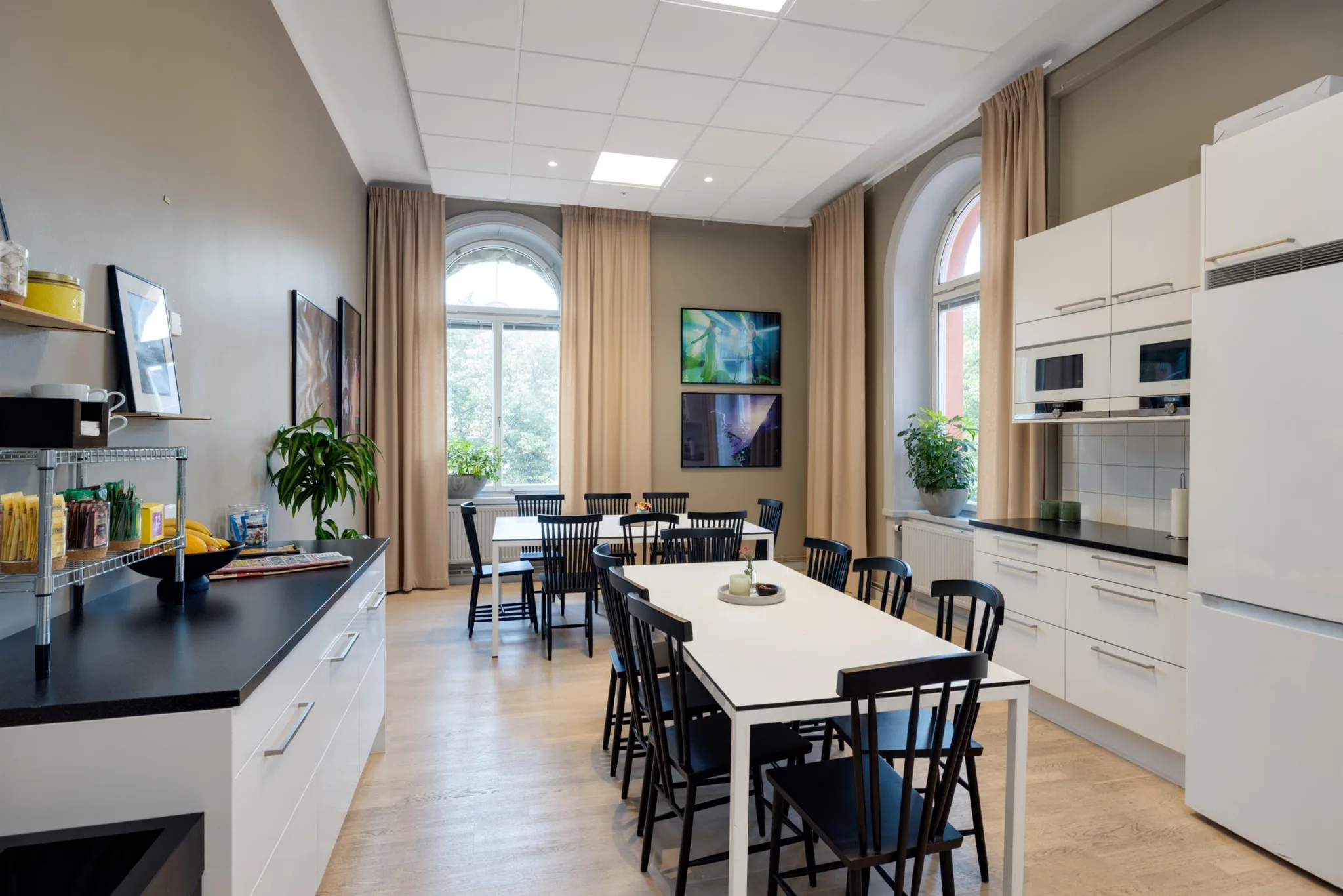 Ett kök med ett långt bord omgivet av svarta stolar, vita skåp, växter och stora fönster med beige gardiner.