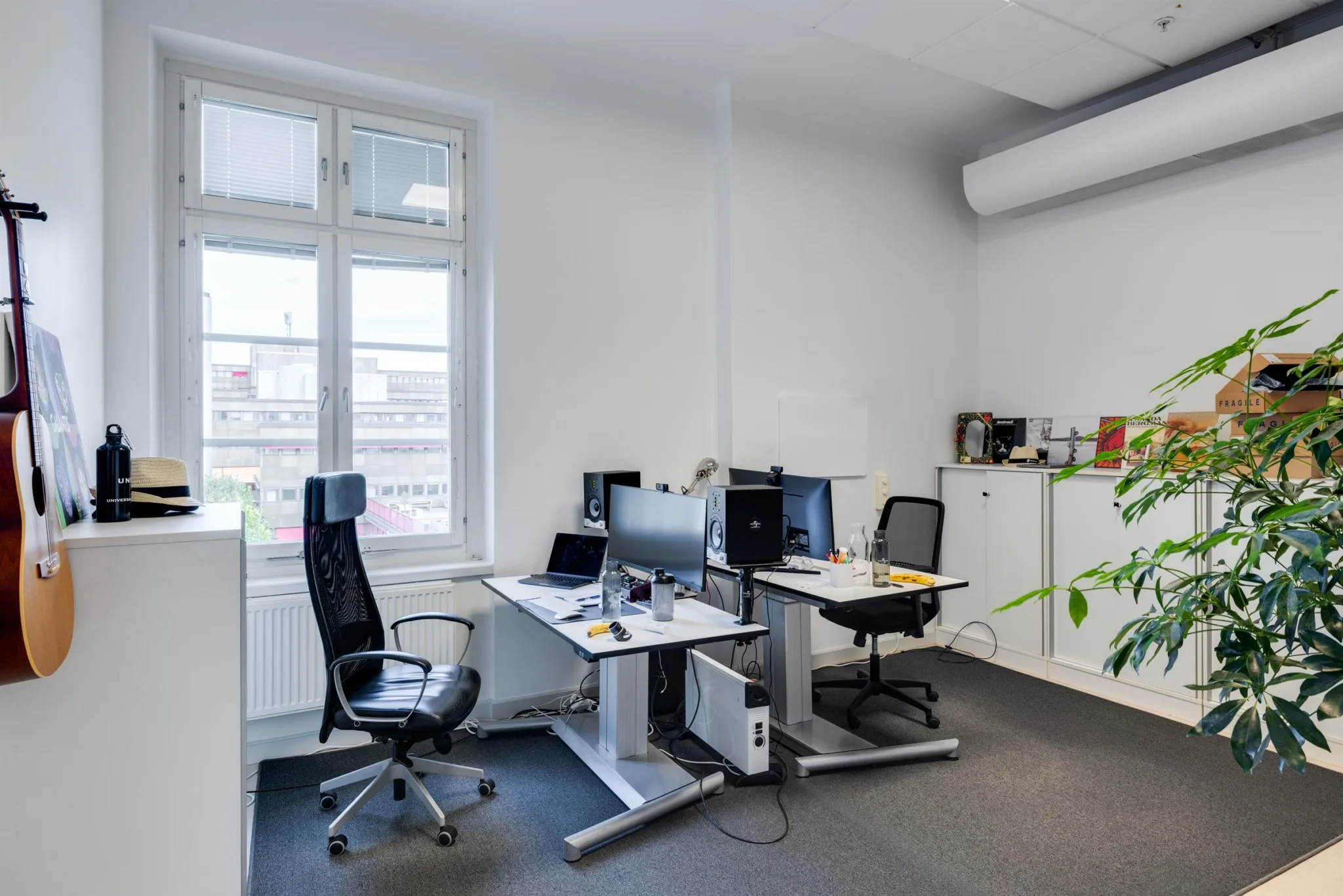 5. Ett enkelt kontorsrum med skrivbord, stolar, datorer och vita skåp nära ett stort fönster.