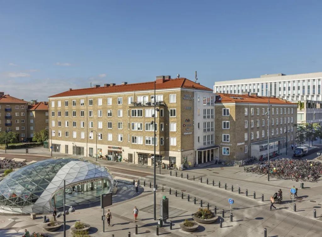 En stadsbild med flera tegelbyggnader, glaskupol och breda trottoarer med gående och cyklar.