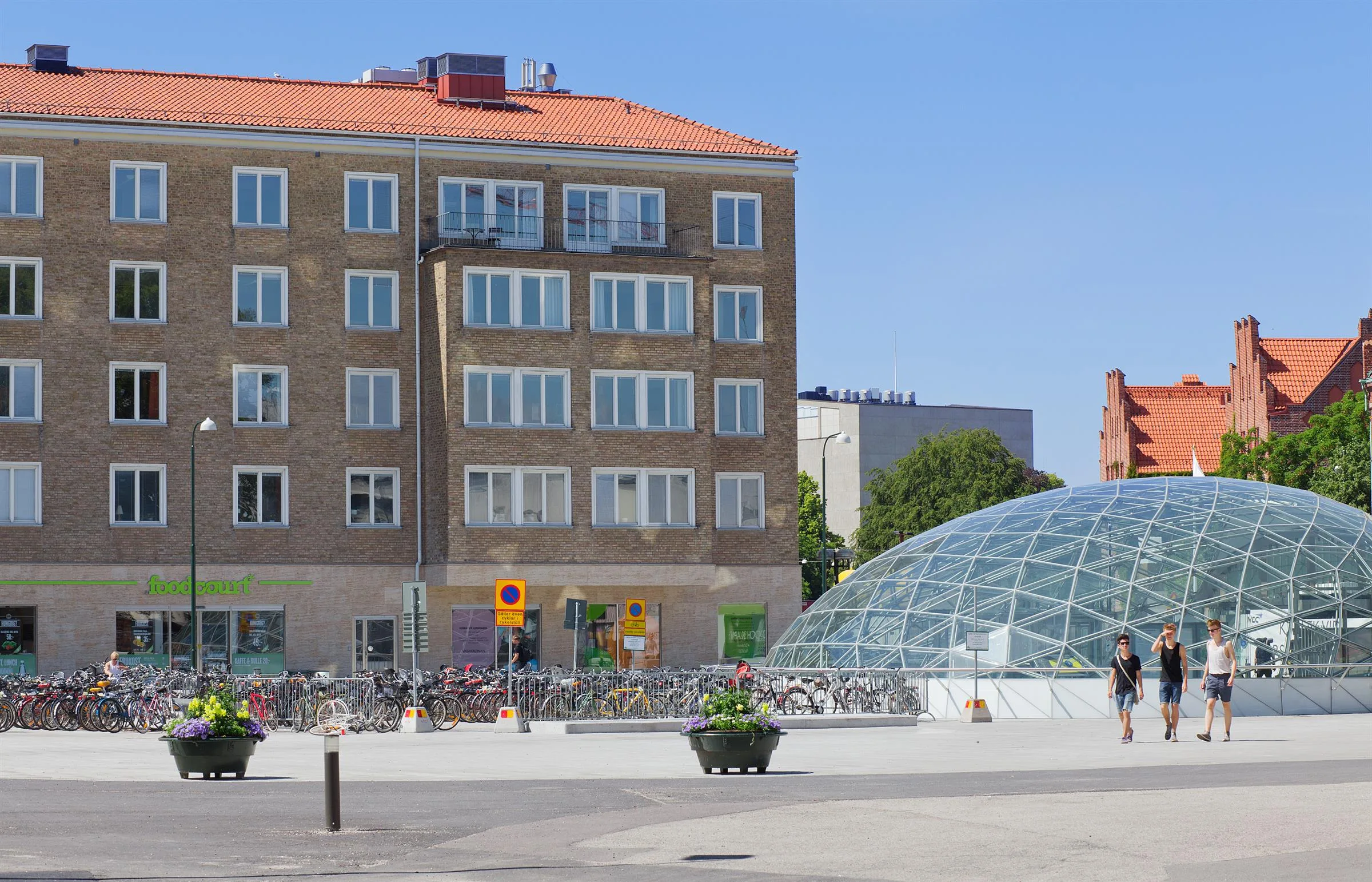 En tegelbyggnad med röda tak och många fönster bredvid en glaskupol, omgiven av cykelställ och gröna växter.