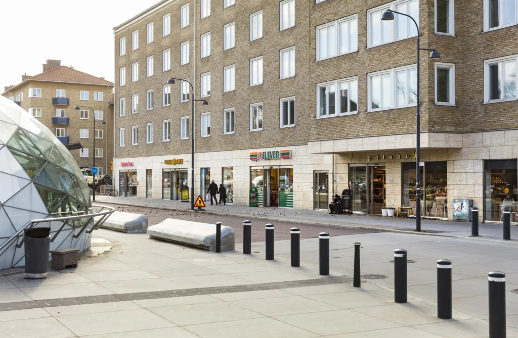 En modern byggnad med butiker på bottenvåningen och en bred trottoar med bänkar och stolpar.
