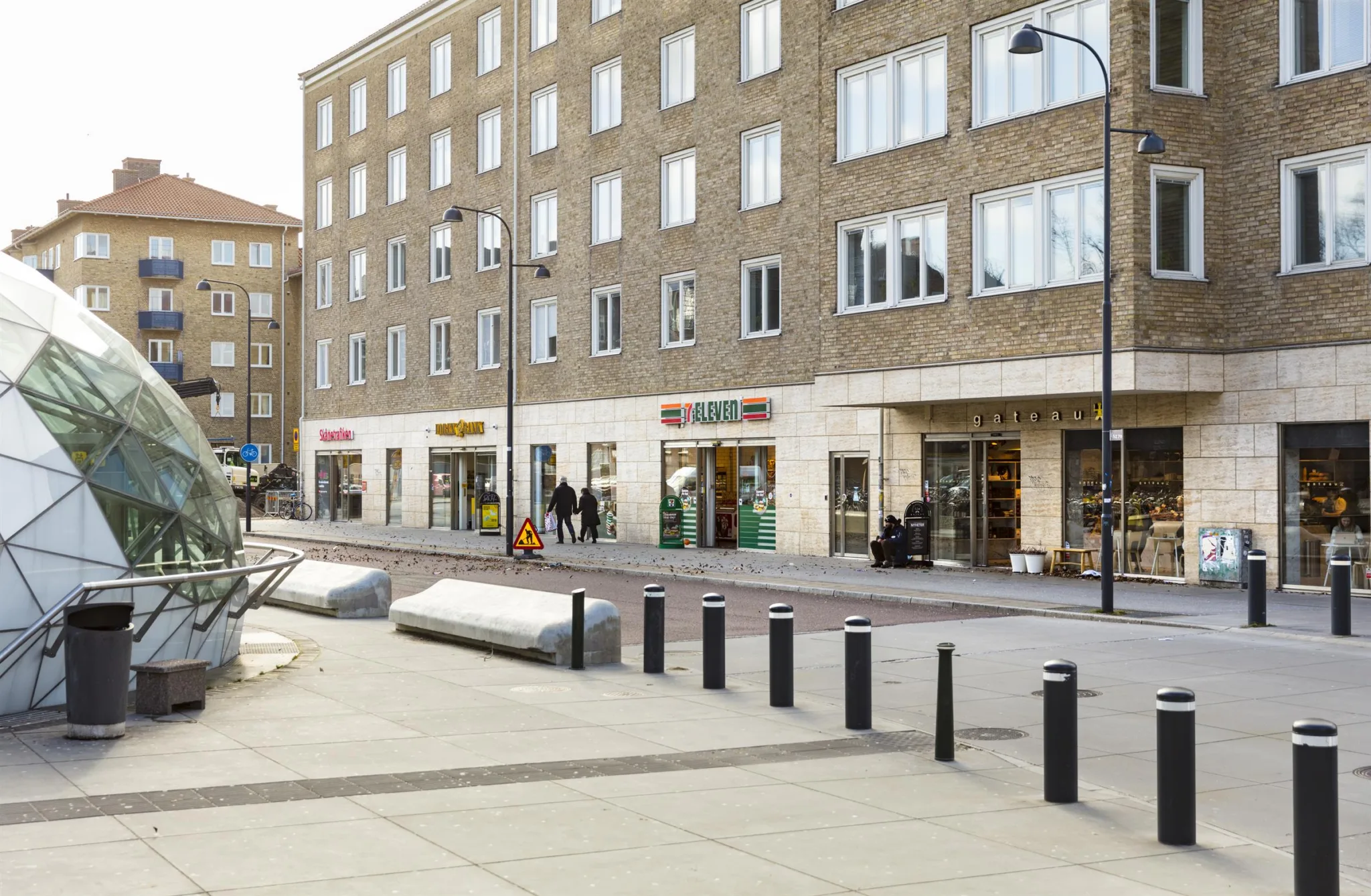 En modern byggnad med butiker på bottenvåningen och en bred trottoar med bänkar och stolpar.