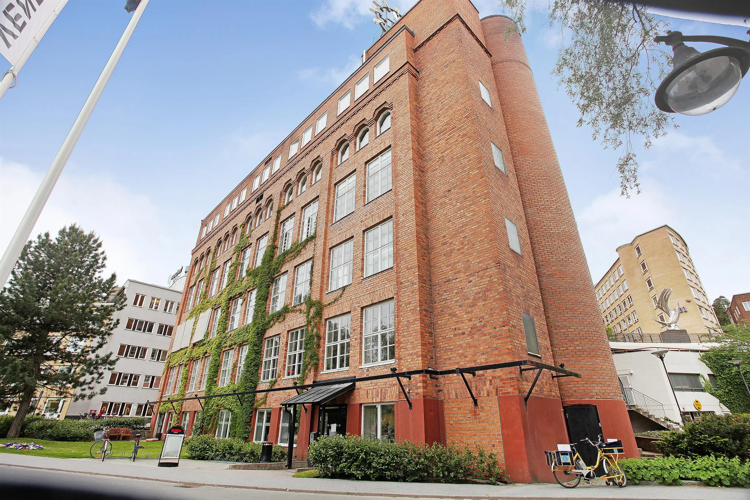 En stor tegelbyggnad med flera våningar, täckt av klätterväxter och med en huvudentré vid trottoaren.