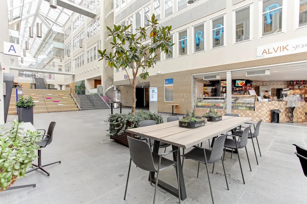Ett öppet atrium med sittplatser vid ett långt bord, växtdekorationer och glasfasader med butiker och kontor.