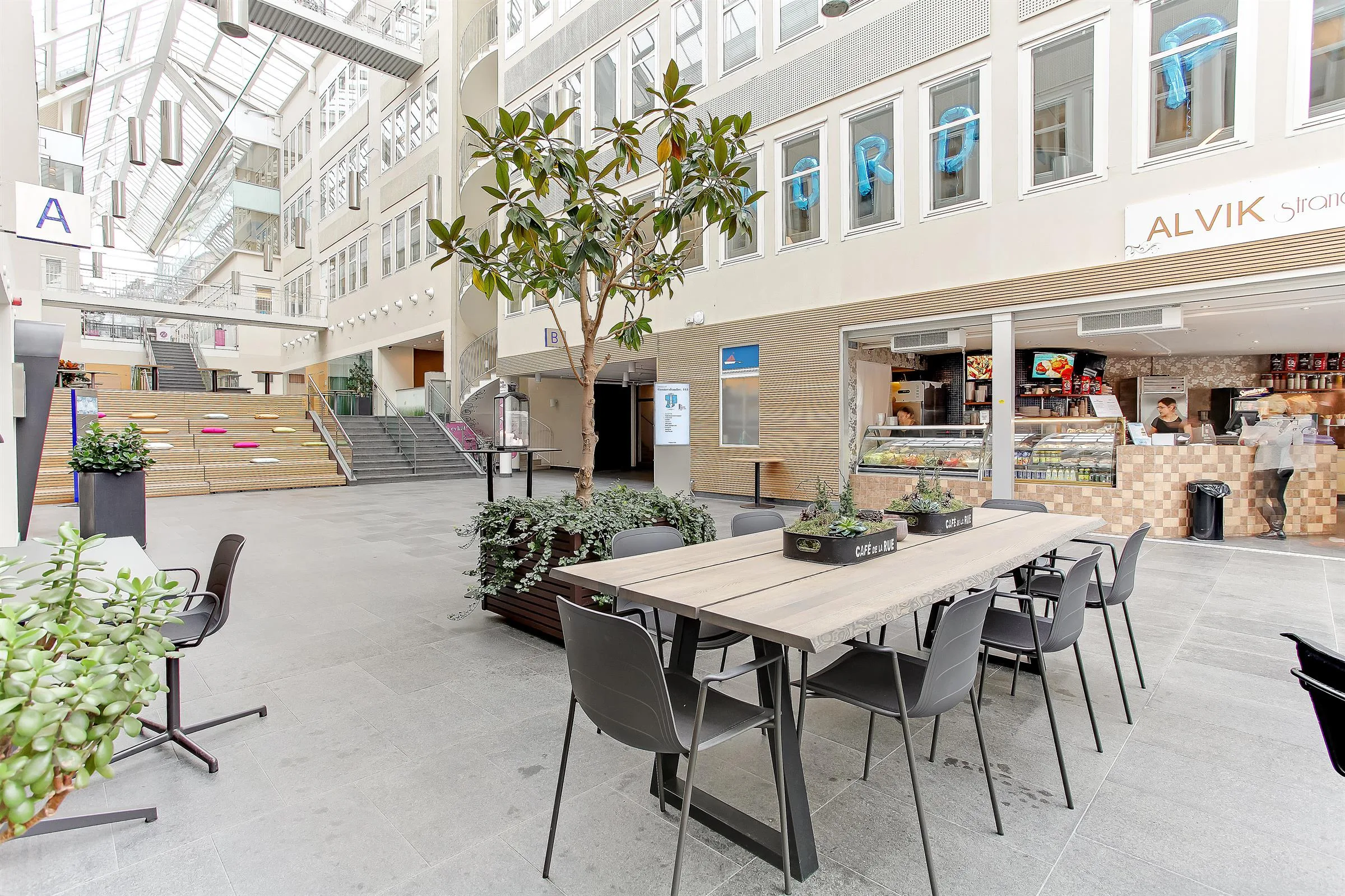 Ett öppet atrium med sittplatser vid ett långt bord, växtdekorationer och glasfasader med butiker och kontor.