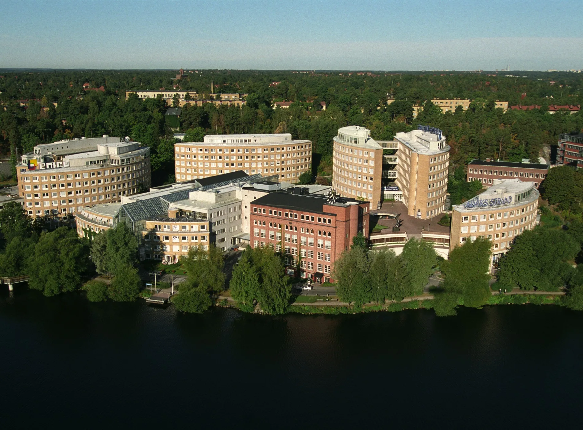 Flygbild av flera byggnader omgivna av träd och vatten.