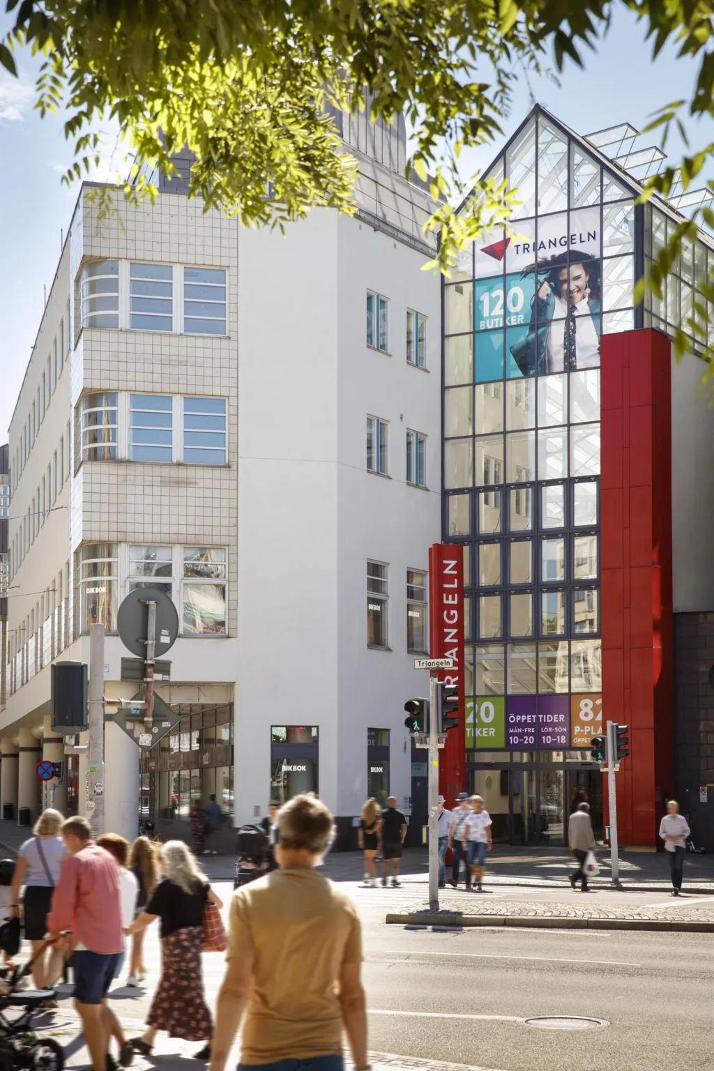 En modern byggnad med glasfasad, röda detaljer och människor som promenerar utanför.