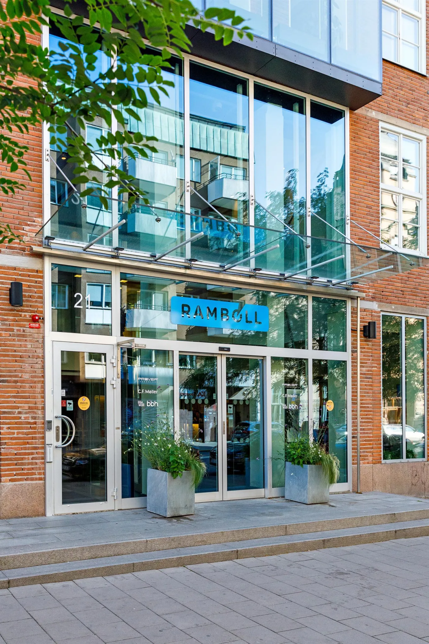 Entré till en byggnad med glasdörrar och texten "RAMPA" ovanför, omgiven av tegelväggar och gröna växter i krukor.