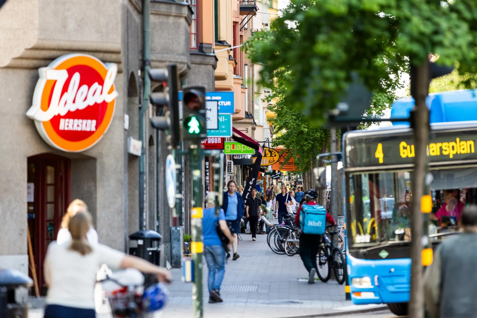 En stadsgata med människor som promenerar, cyklar och en blå buss som närmar sig, med en skylt för "Real" synlig till vänster.