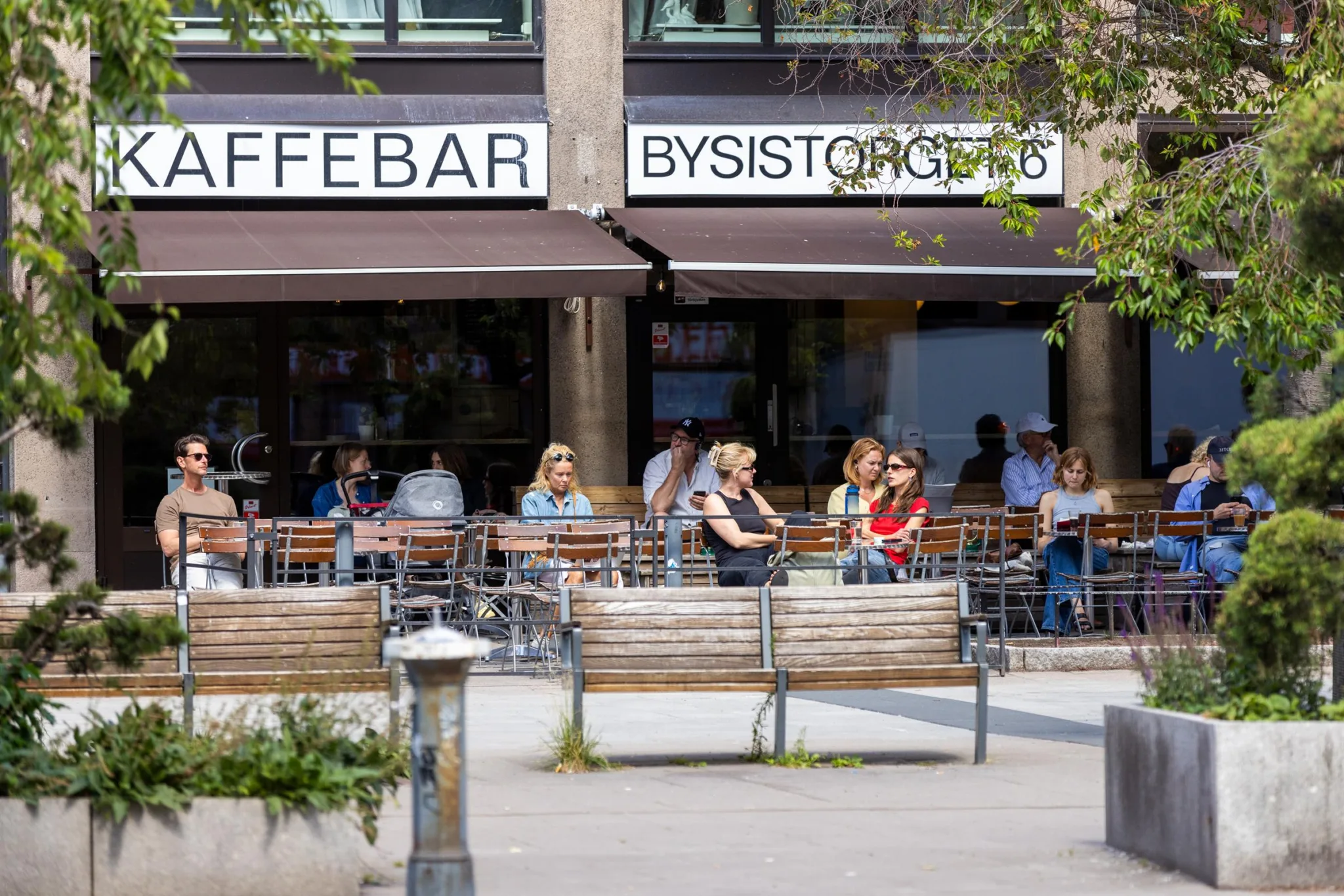 Ett kafé med uteservering där människor sitter på bänkar och njuter av solen, texten ”Kaffebar Bysistorgets” syns på markisen.