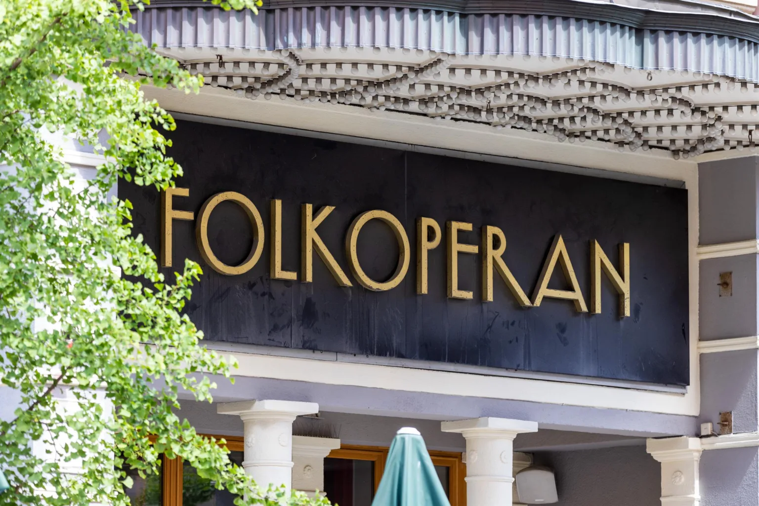 En fasad med texten "FOLKOPERAN" i guldfärg på en svart skylt, omgiven av trädgrenar.