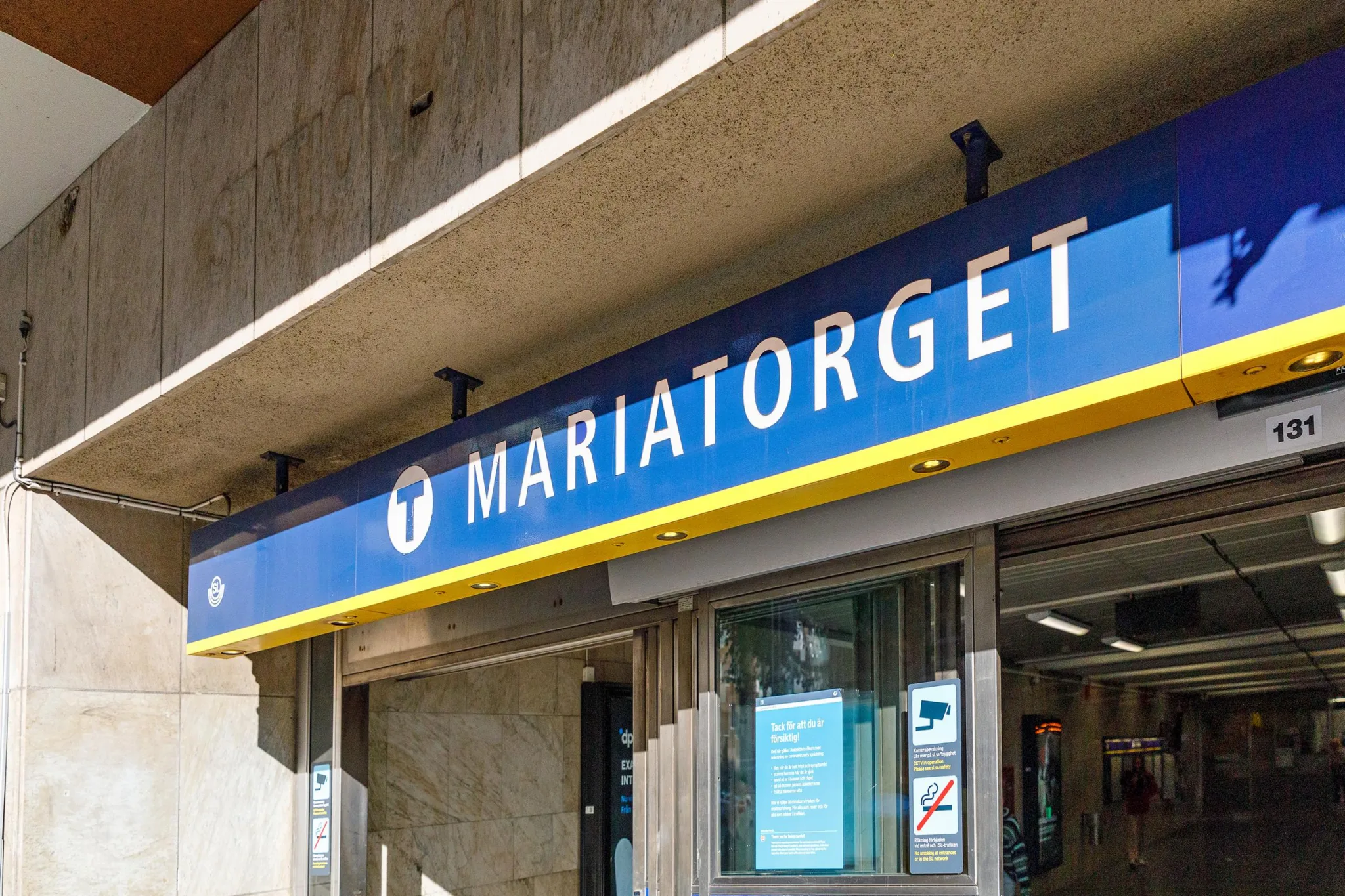 En skylt för tunnelbanestationen Mariatorget, med blå bakgrund och vit text, monterad på en betongvägg ovanför ingången.
