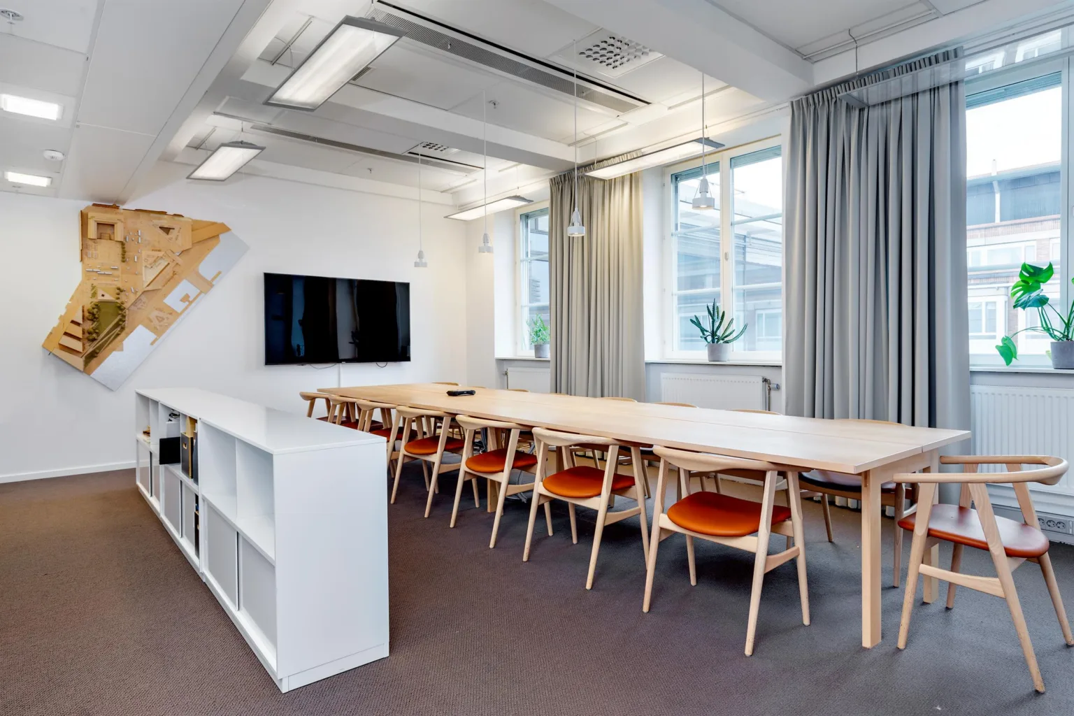 Ett konferensrum med långa träbord, stolar med orangea dynor, en TV på väggen och stora fönster.
