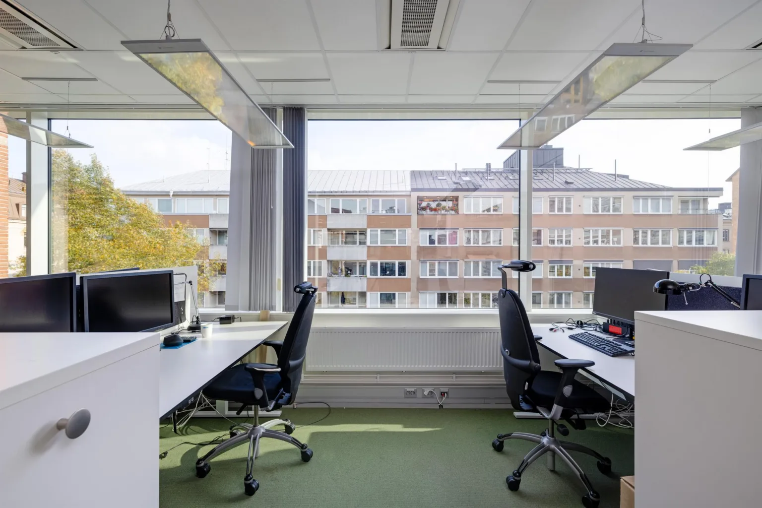 Ett modernt kontorsutrymme med skrivbord, stolar och datorskärmar, med en stor fönstervy mot byggnader och träd.