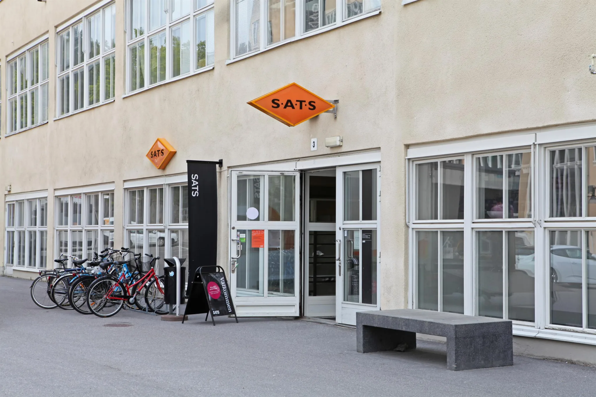 En byggnadsentré med glasdörrar och SATS-logotypen på orangea skyltar, cyklar parkerade bredvid.