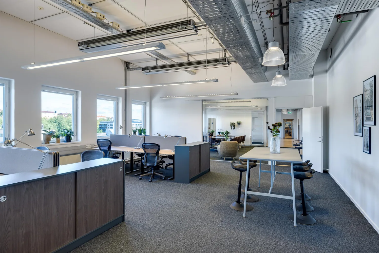 En modern kontorslokal med arbetsstationer, ståbord och synlig ventilation i taket.