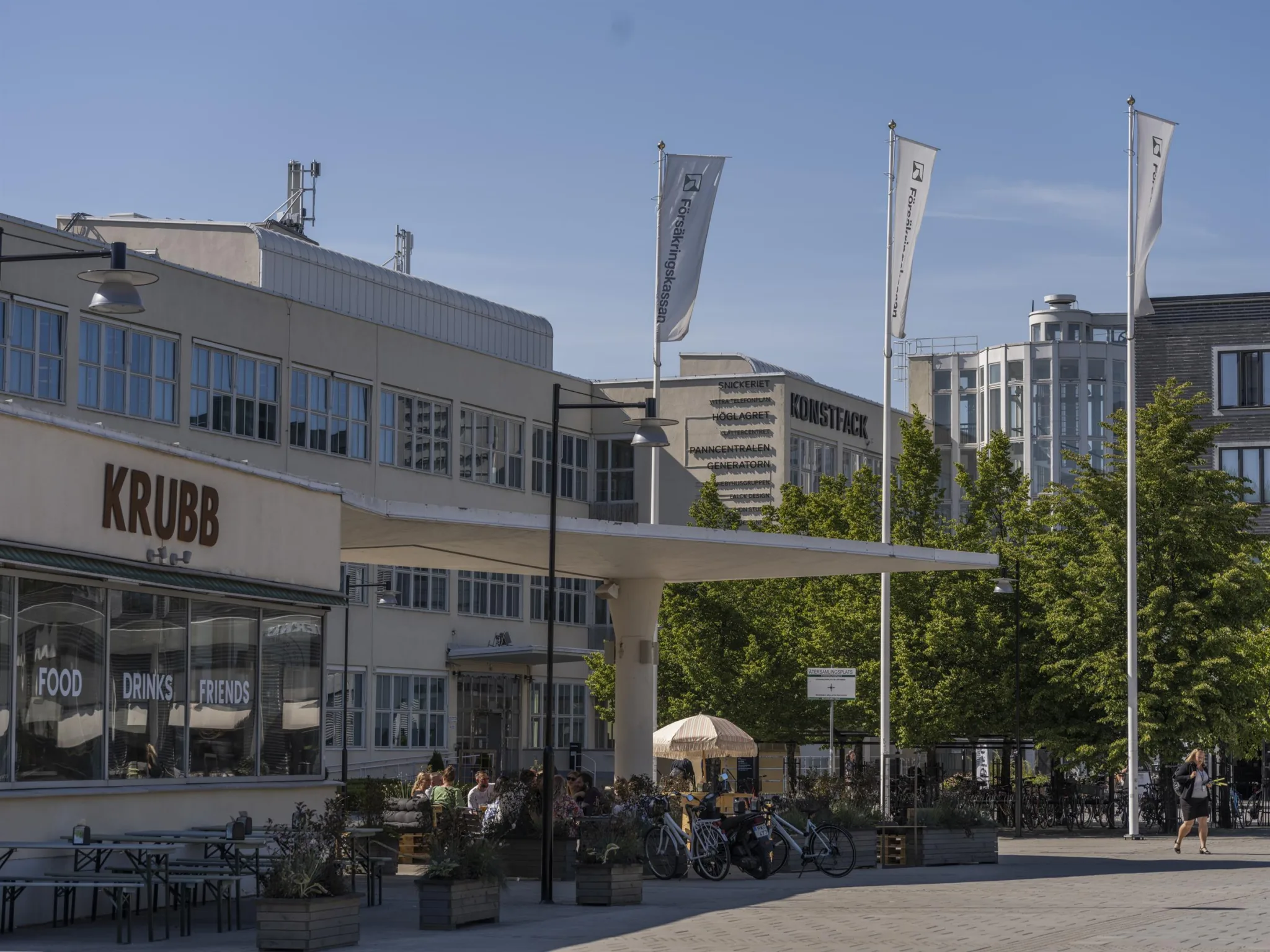 En utomhusbild av byggnader med caféer och sittplatser, omgivna av träd och flaggor under en klarblå himmel.