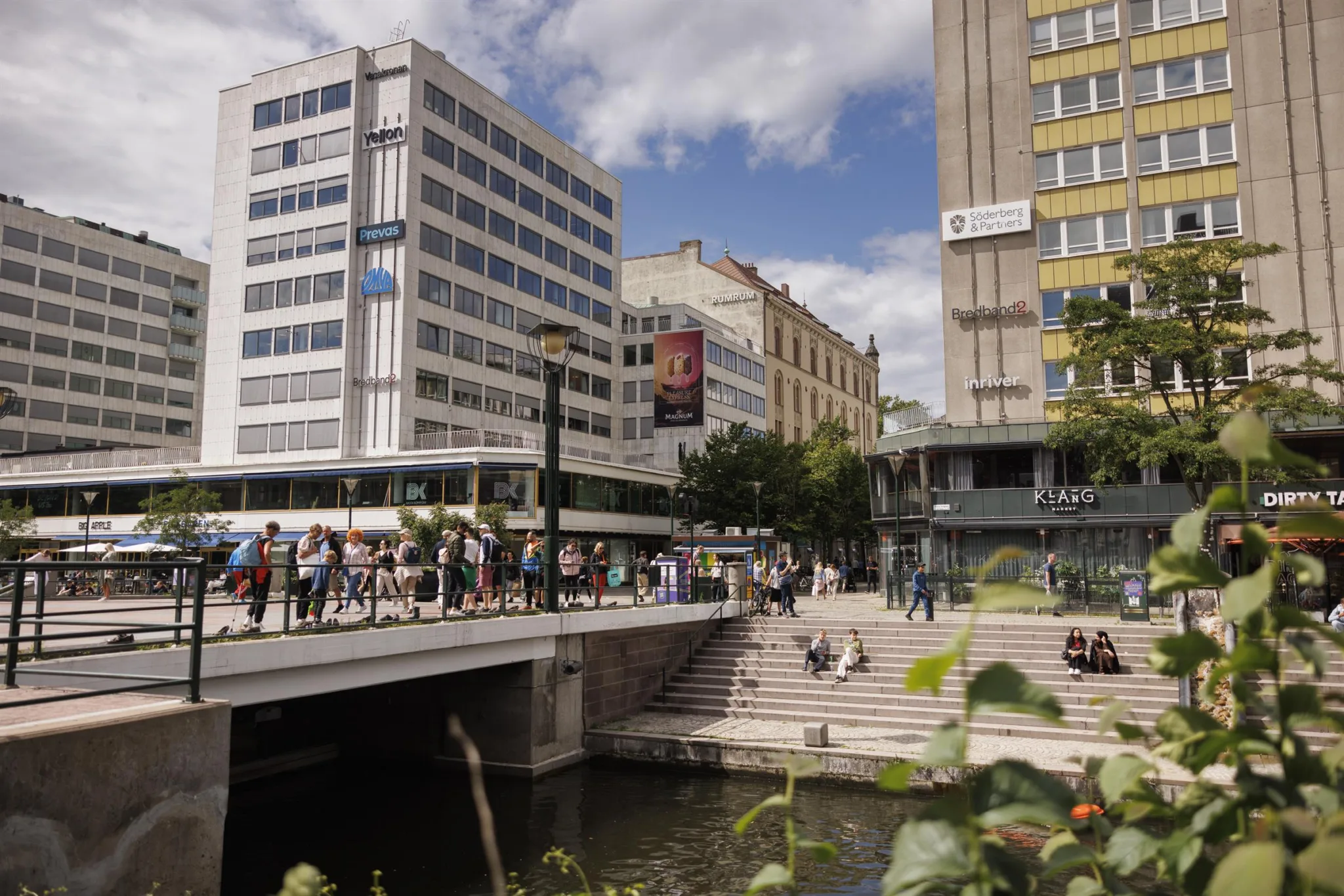 En stadsmiljö med moderna byggnader, människor som promenerar och en kanal med grönska.