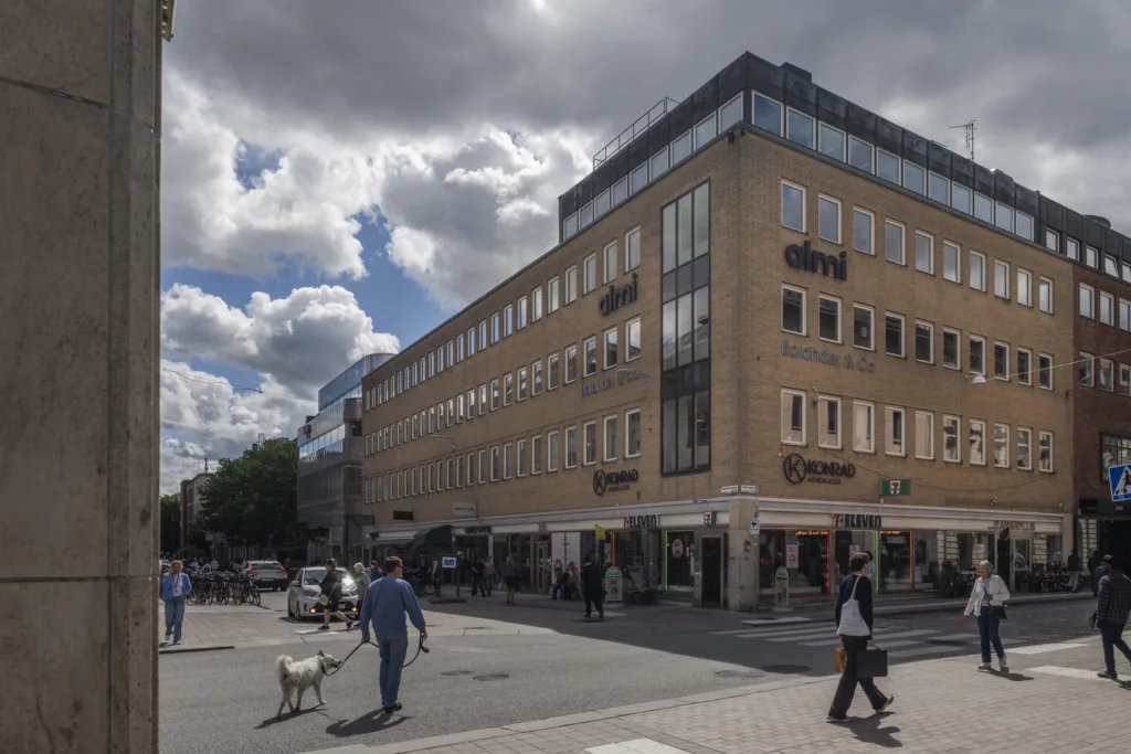 En modern byggnad med en gul fasad vid ett hörn; människor och en hund promenerar under molnig himmel.