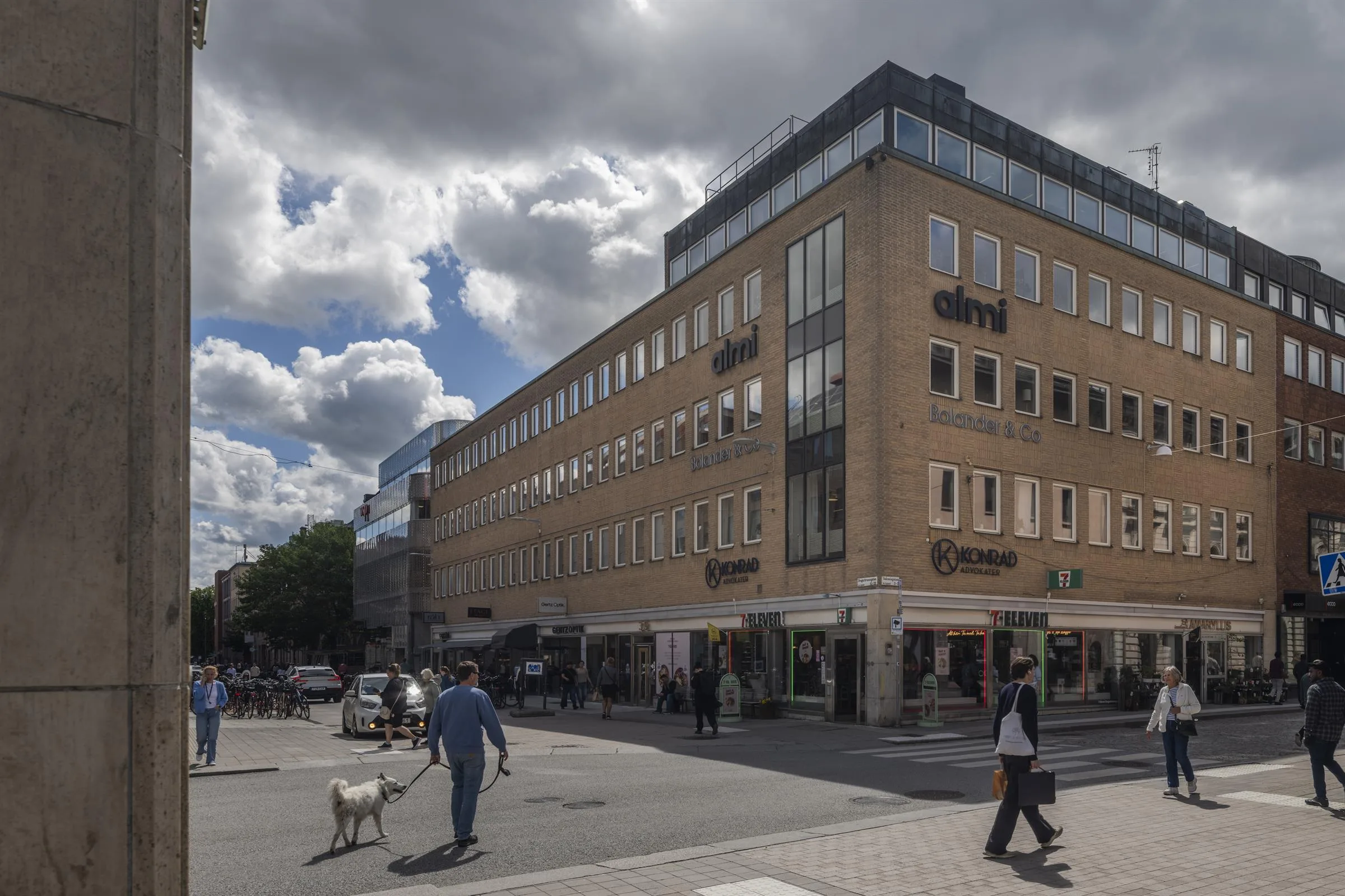 En modern byggnad med en gul fasad vid ett hörn; människor och en hund promenerar under molnig himmel.
