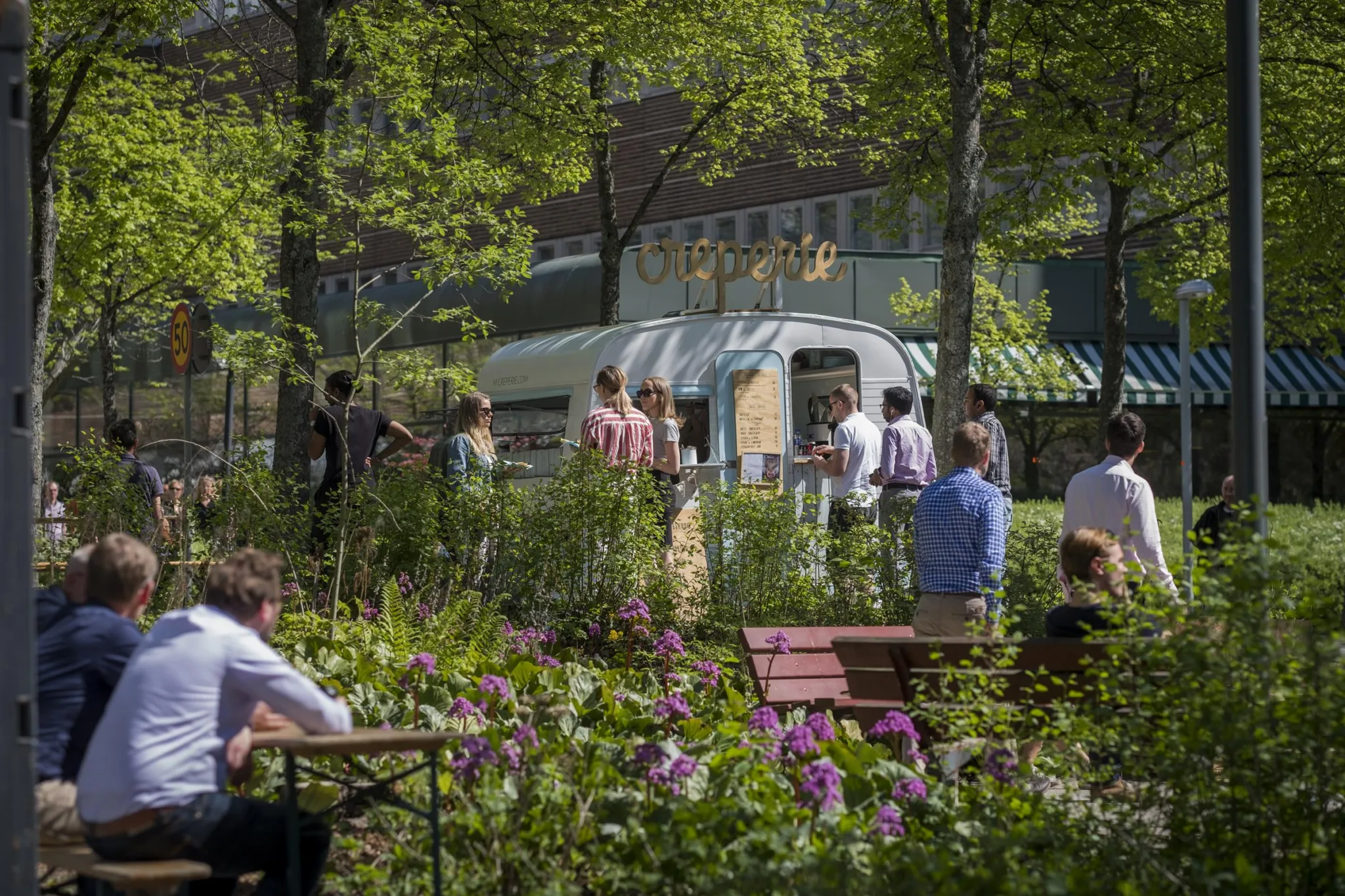 2. En uteplats med människor som köar vid en matvagn, omgiven av grönska och blommor.