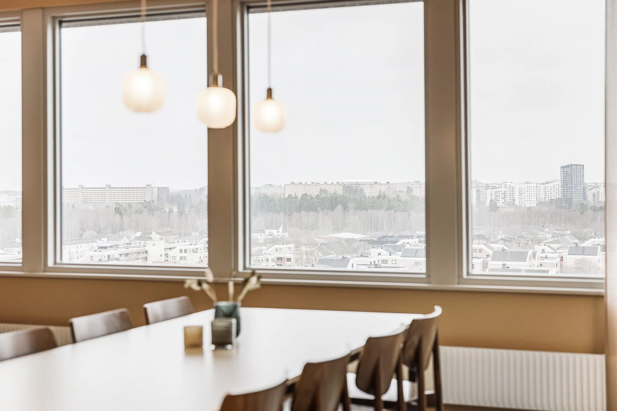 1. Ett ljust mötesrum med stora fönster, en lång vit bordsskiva och bruna stolar, med tre hängande lampor ovanför.