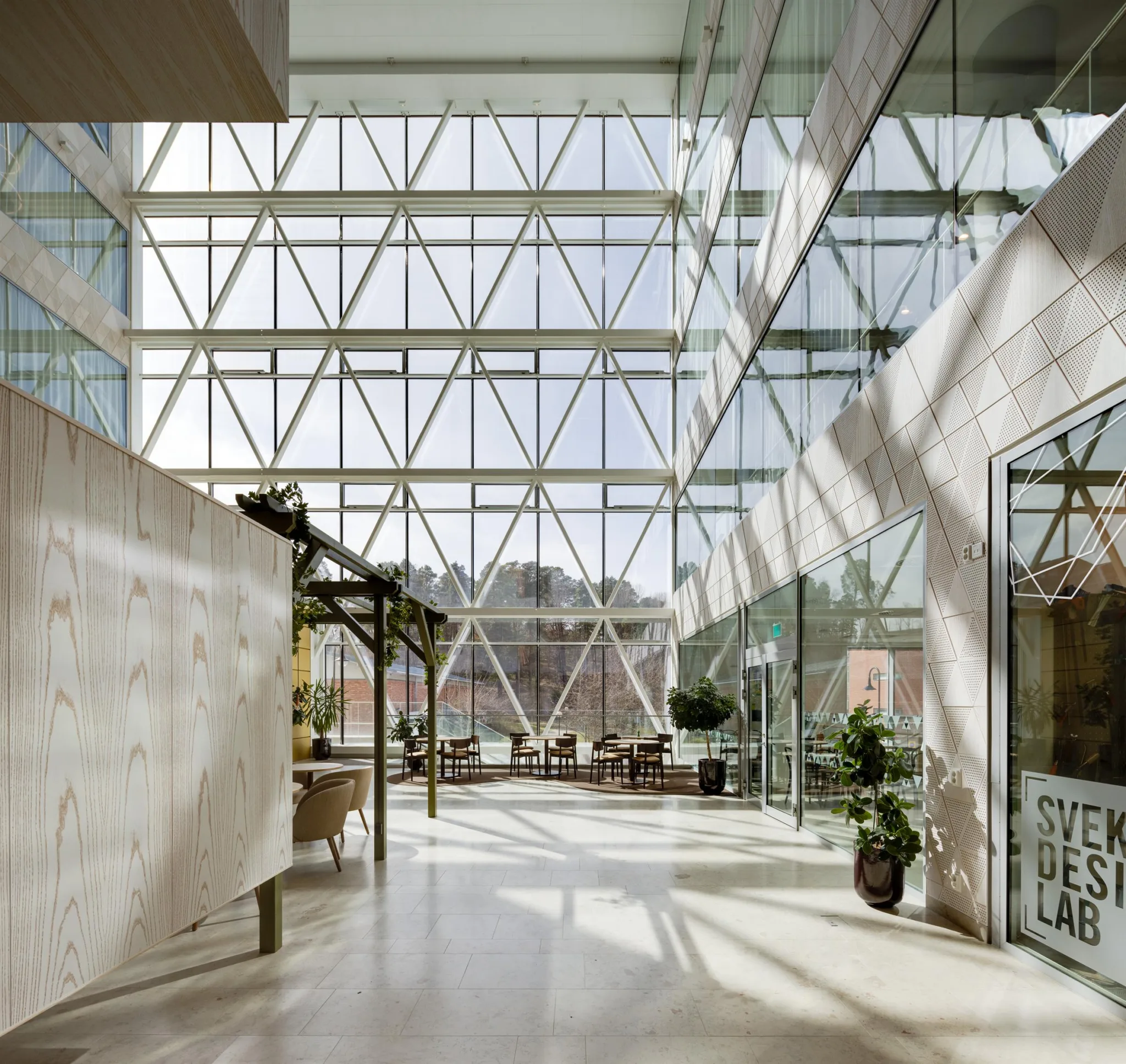 Ett ljust atrium med stora glasväggar, grönska och sittplatser.
