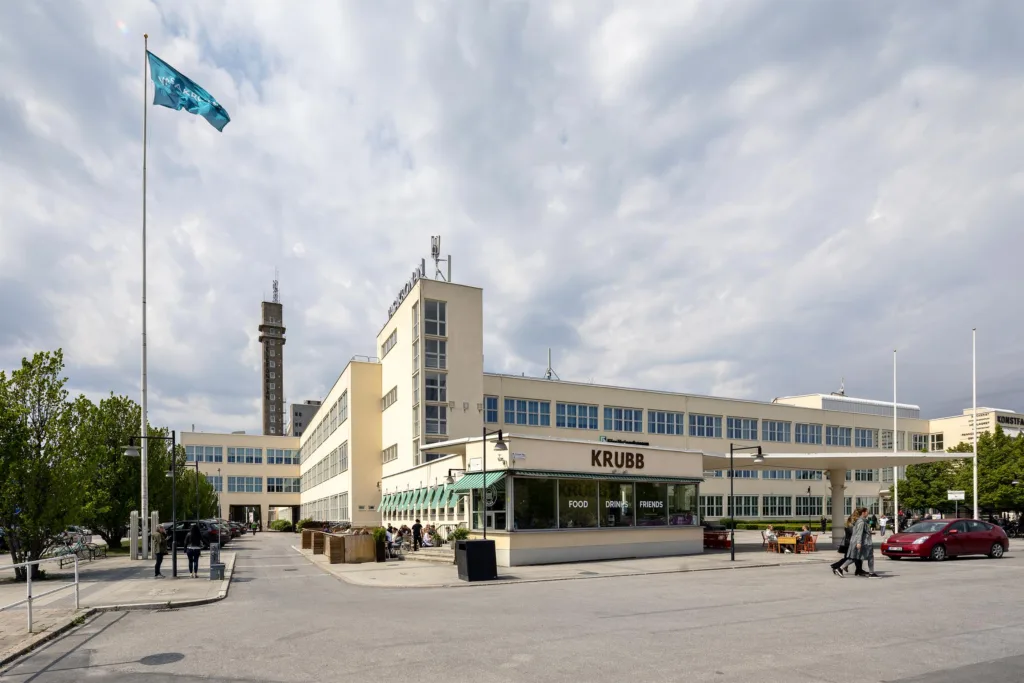 En vit byggnad med flera våningar och flagga på vänster sida, med en tornstruktur i bakgrunden under molnig himmel.