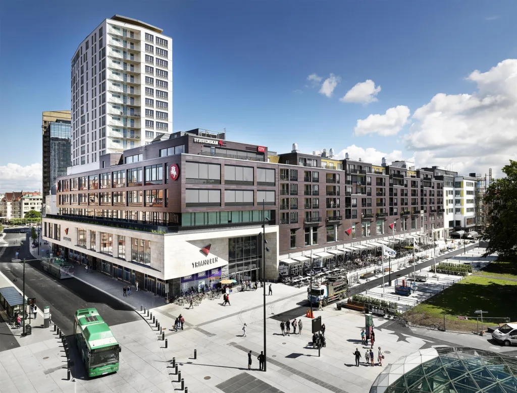 Ett stadstorg med flera moderna byggnader, en spårvagn och människor som promenerar.