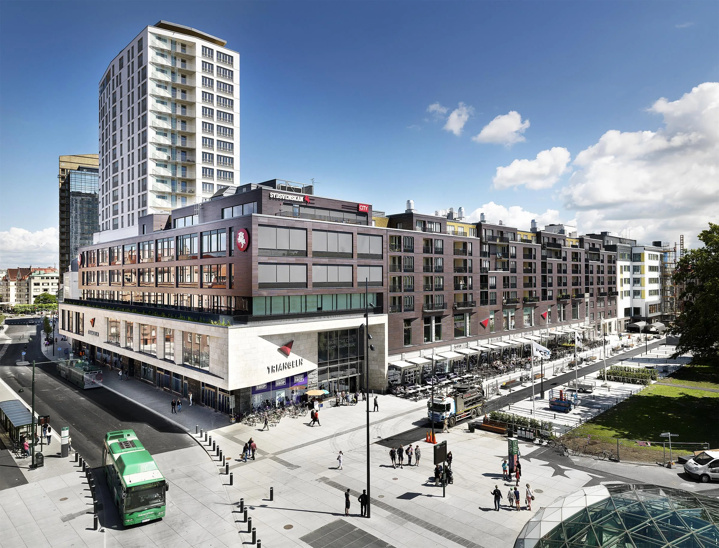 Ett stadstorg med flera moderna byggnader, en spårvagn och människor som promenerar.