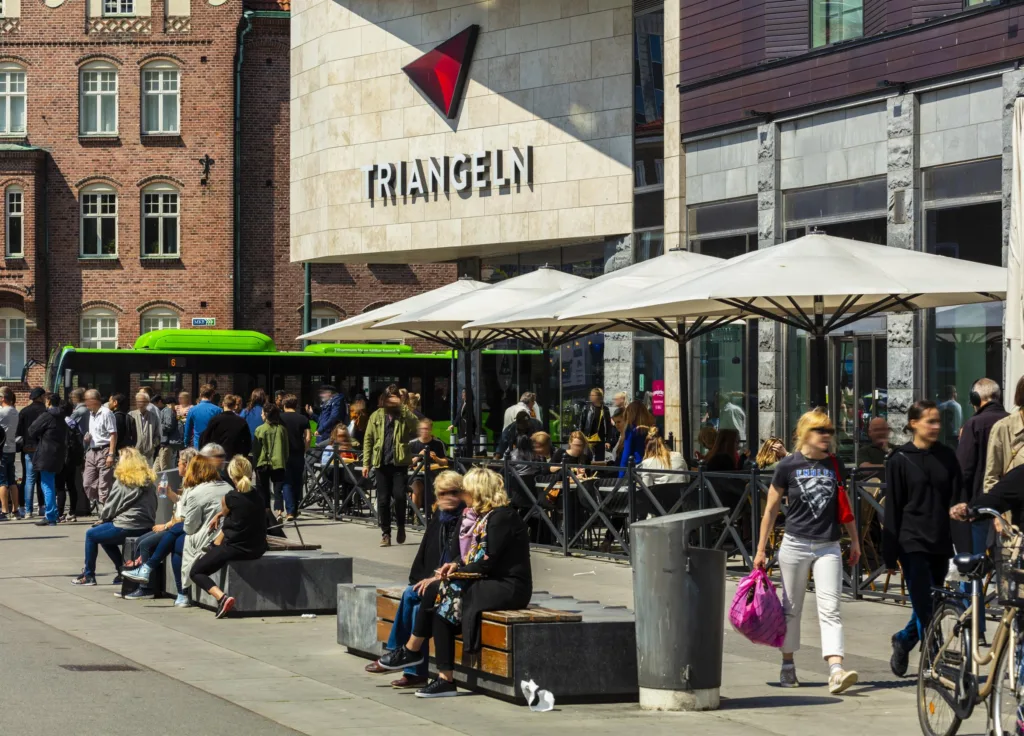 Personer sitter och promenerar framför Triangelns köpcenter, med byggnaden och skyltar tydligt synliga.