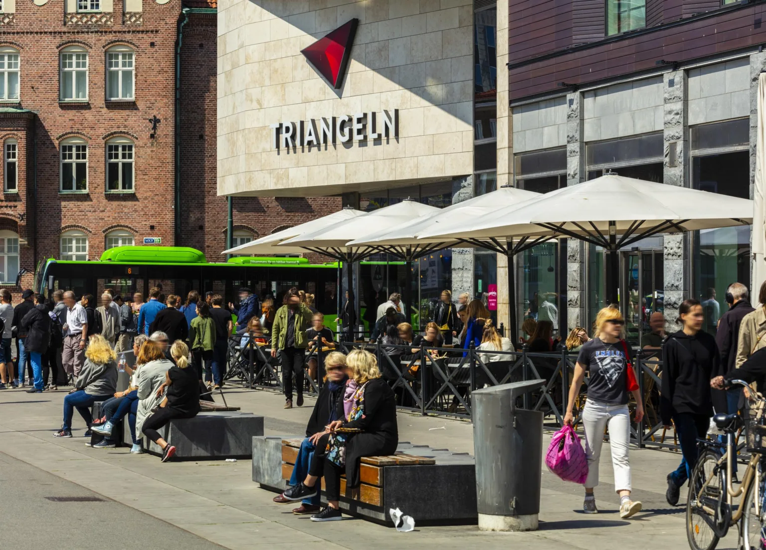 Personer sitter och promenerar framför Triangelns köpcenter, med byggnaden och skyltar tydligt synliga.