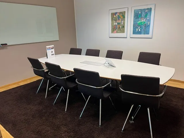 Ett konferensrum med ett vitt bord omgiven av åtta svarta stolar, en whiteboard på väggen och två färgglada tavlor.