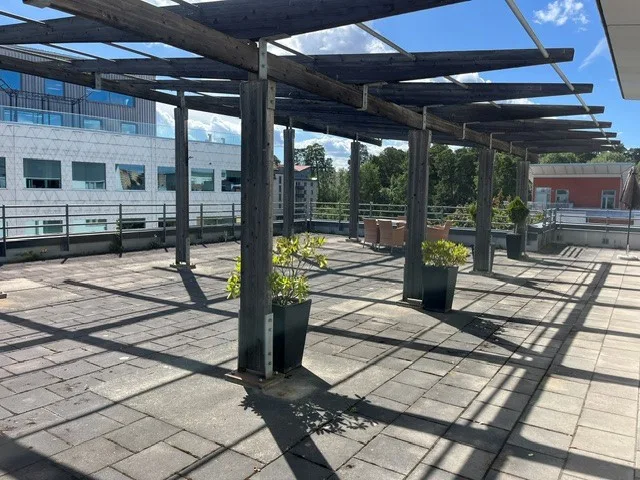 En utomhusterrass med pergola, stenbeläggning och gröna växter i krukor under klarblå himmel.
