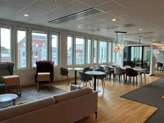 En lounge med soffor, fåtölj, bord och stora fönster med utsikt över stadsmiljö.