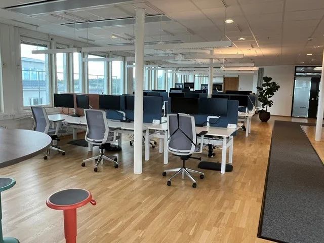 Ett öppet kontorslandskap med skrivbord, kontorsstolar, datorer och stora fönster som ger mycket ljusinsläpp.