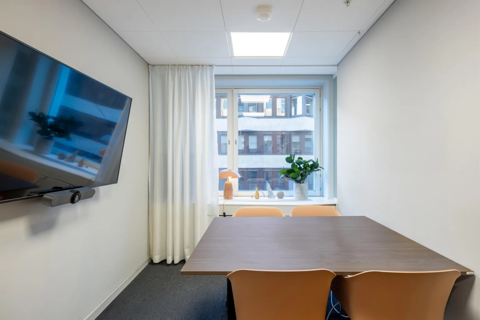 2. Ett mötesrum med bord, stolar, en väggmonterad skärm och ett fönster med utsikt över en byggnad.