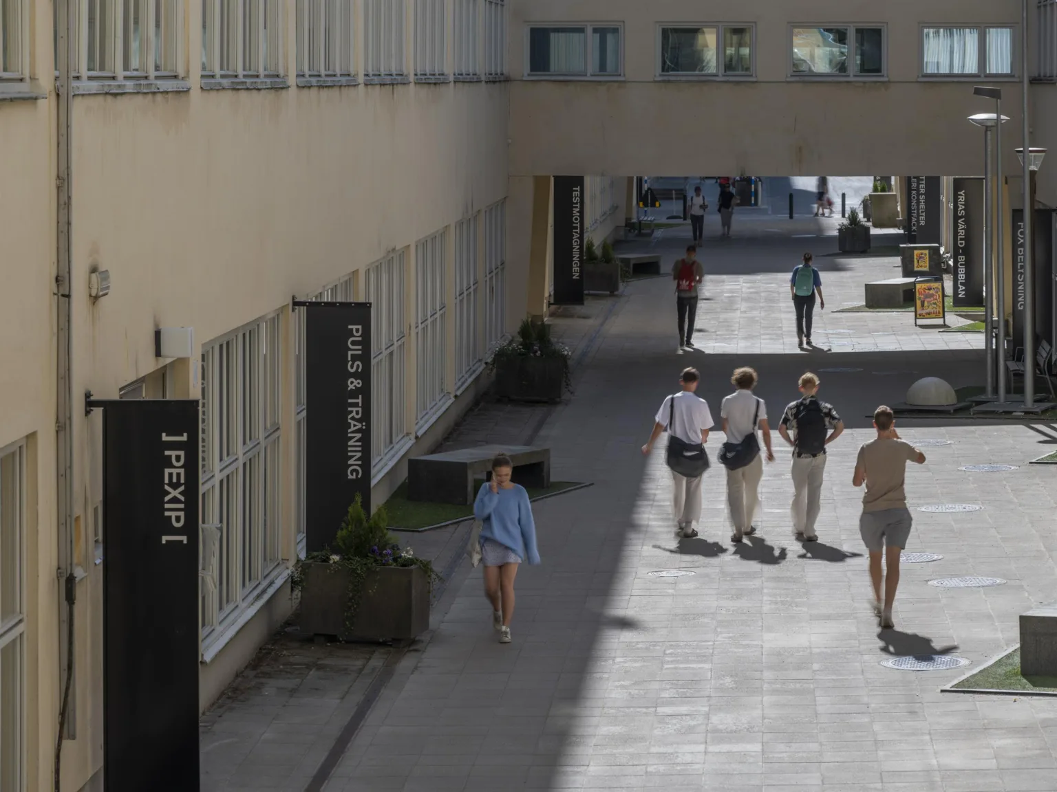 En gångväg mellan byggnader med människor som promenerar, en skylt med texten "1 trappa" synlig på en vägg.