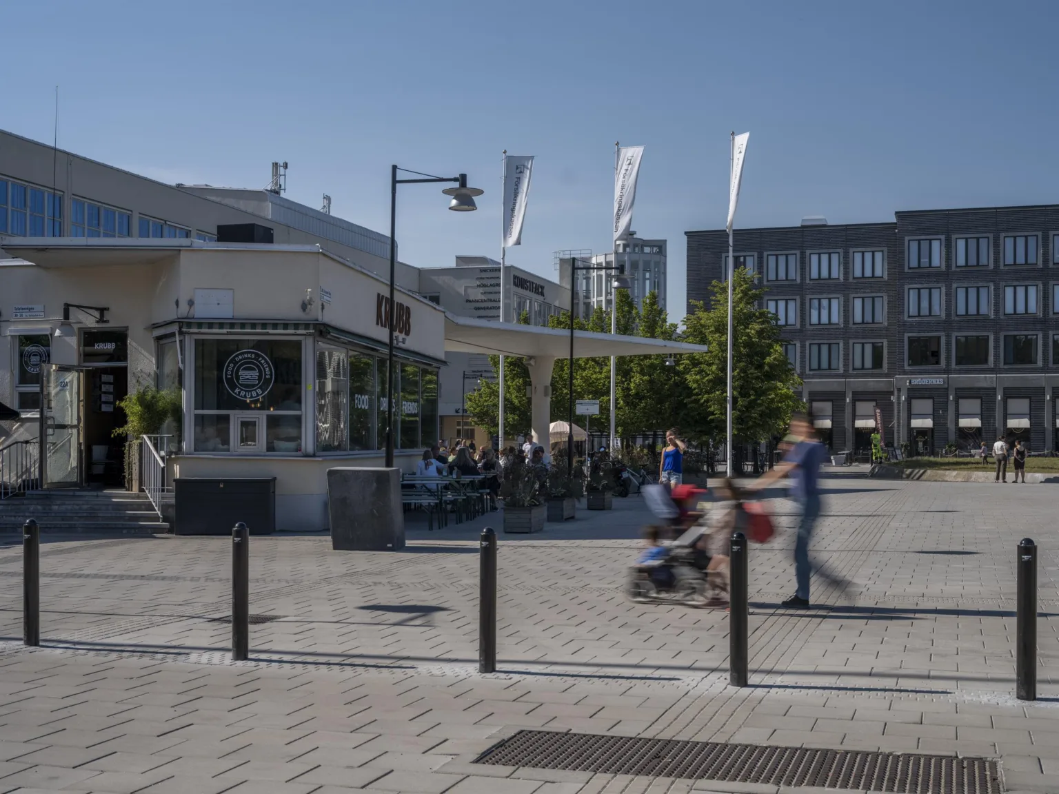 En stadsmiljö med människor som går, en uteservering, byggnader och flaggor i bakgrunden.